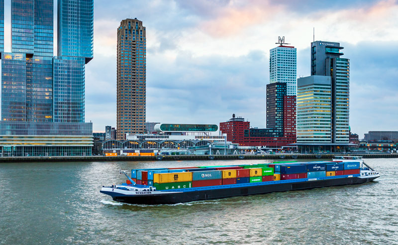 Huge container ship full of freight boxes cruising the river with Rotterdams skyline in the background.