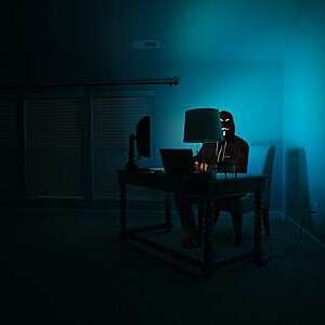 Hacker dressed in black and wearing a Guy Fawkes mask sits at a desk in a dark room, illuminated only up by the dim light of his laptop screen.