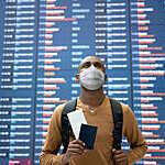 Portrait of a black male traveler wearing a facemask at the airport with the flight schedule at the background while holding his passport - travel concepts